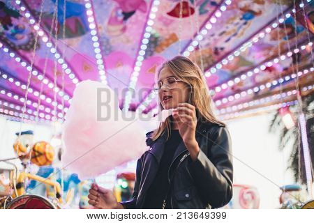Fashionable young woman in black trendy leather jacket stands in middle of county fair next to attraction rides or carousel holds big pink cotton candy or sugar floss enjoys date night or holidays