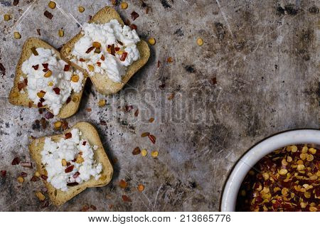 Flat lay photograph of miniature toast topped with white cheese and red pepper flakes sitting on an old metal pan background.