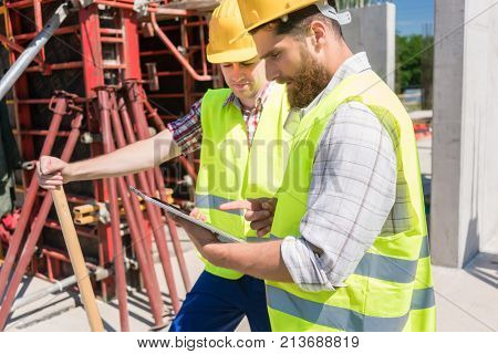 Two young construction workers smiling while using a tablet for online communication through social media during break at work