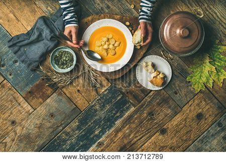 Flat-lay of female hands and Fall warming pumpkin cream soup palte with croutons and seeds over rustic wooden table background, top view. Autumn vegetarian, vegan, healthy comfort food eating concept.