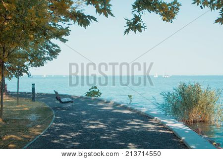 A view of Balaton lake with white yachts at the horizon and trees bench and footpath at the foreground on sunny autumn day Tihany Hungary.