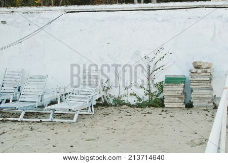 Old White Vintage Wooden Sun Loungers Standing Near The Wall At The Beach After The End Of A Summen