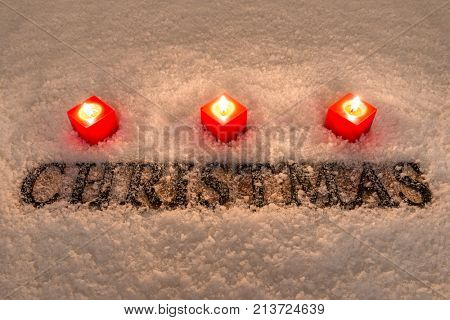 The word Christmas in wooden letters on a snow background illuminated by three red candles.