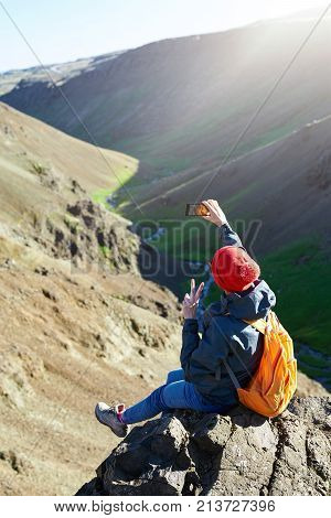 back view of woman traveler on a walk in the Valley of the river of Hveragerdi Iceland. Hiking Tour of Reykjadalur Hot Springs. woman sits on the cliff and use smartphone making selfie