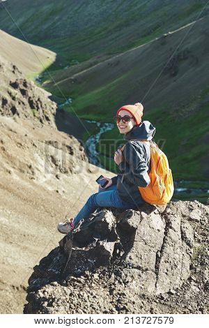 back view of woman traveler on a walk in the Valley of the river of Hveragerdi Iceland. Hiking Tour of Reykjadalur Hot Springs. woman sits on the cliff and use smartphone