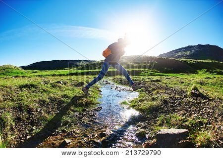 woman traveler with small orange backpack on a walk in the Valley of the river of Hveragerdi Iceland. Hiking Tour of Reykjadalur Hot Springs. woman jumping over creek