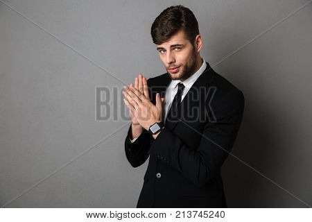 Close-up photo of handsome young man in black suit clap in hands, lookig at camera, isolated over gray background