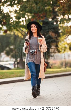 Full lenth photo of pretty modern asian girl holding smartphone and coffee to-go while strolling in public park