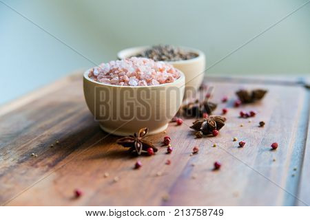 Pink Himalayan salt and coriander seeds in white bowls on wooden surface covered with anise and red peppercorn