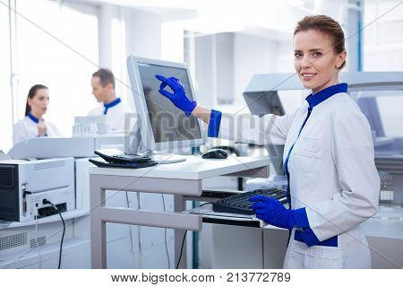 Patient welfare.  Kind professional female researcher  gazing straight while pointing at the screen and posing in the lab