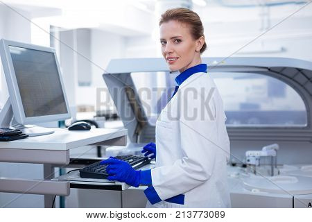 Science in lab. Attractive kind female laboratorian entering data waiting for other results while posing near the medical equipment