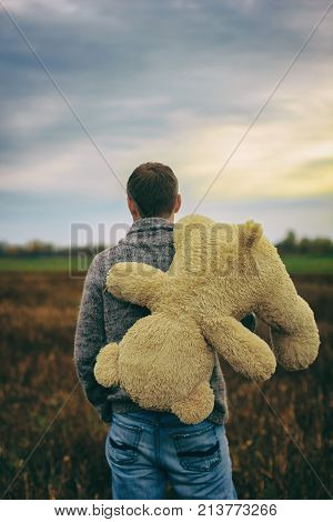 Man holding teddy bear behind him. He walks the field and looks out into the distance