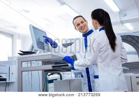 Flirting at work. Handsome male scientist  holding his hand near the screen and   flirting with female scientist  while she standing with her back