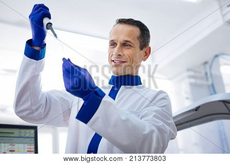 Good perspective. Low angle of pleasant smart laboratory assistant  holding glassware looking at it and smiling
