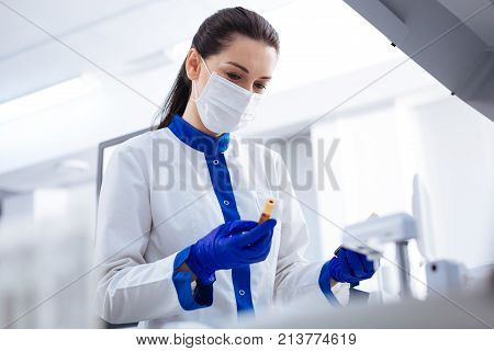 Time for analyses. Brunette serious laboratory assistant holding blood sample for checking on HIV and wearing medical mask