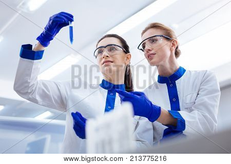 Predicting results. Brunette curious scientist  bringing up test tube while blond beautiful  female scientist pointing her finger and telling details