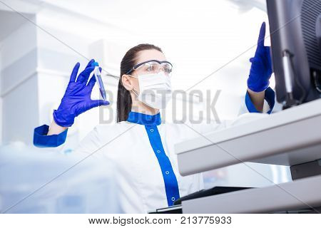 Unknown  results. Concerned attentive young lab assistant  has test tube in her hand  touching the screen while wearing medical mask