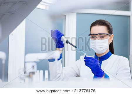 Power in drop. Brunette tranquil laboratory assistant  dropping liquid into glassware which she carrying in the hand while looking down