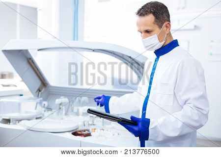 Cutting edge equipment. Attractive focused  male scientist reading instruction for innovative centrifuge machine  while holding tablet and standing in lab