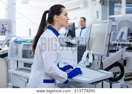 Nice touch. Energetic positive female laboratory assistant looking at the screen, typing while standing  near the medical equipment