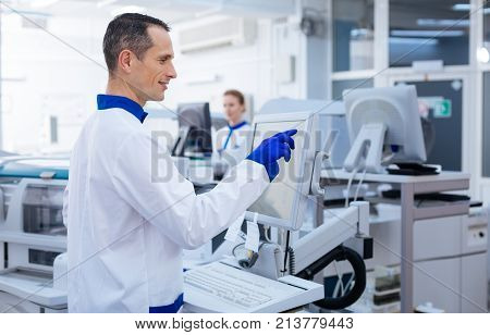 Successful experiment. Happy pleased male researcher achieving necessary reaction while placing his hand on the keyboard and brining up  glassware while standing in the lab