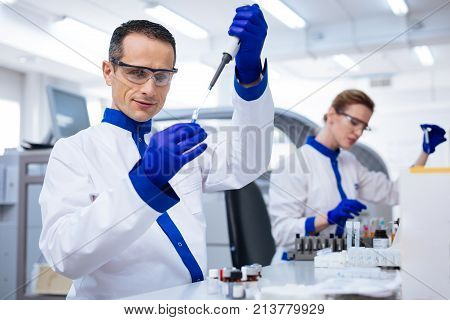 Routine test. Inspired prominent male laboratory assistant holding the glassware and dropping reagent there while wearing medical glasses