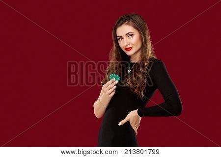 Pretty long hair woman in black dress holding chips for gambling in casino. Studio shot on red background. Poker. Two green Chips in Hand