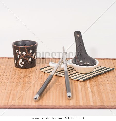 Metal chopsticks on ceramic chopsticks rest.  black porcelan spoon with white spoon rest and black sake glass on bamboo mat. White background. Front view