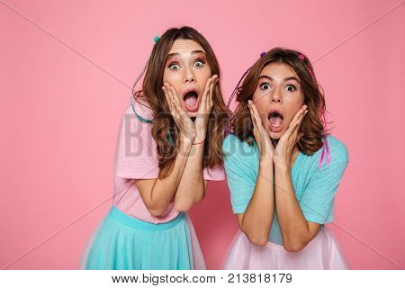 Close-up portrait of two shocked girls in children's clothes touching their cheeks, looking at camera, iolated on pink background