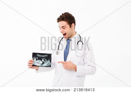 Portrait of a happy young male doctor with stethoscope dressed in uniform pointing finger at x-ray photograph isolated over white background