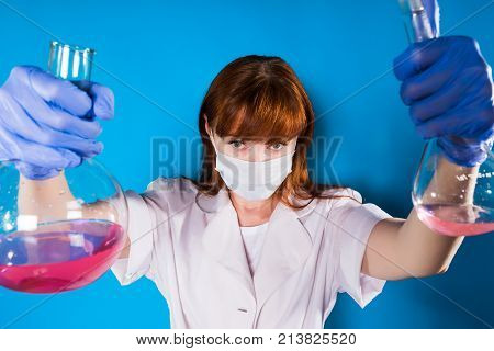an adult red-haired woman chemist in a face mask and a dressing gown is holding albes with chemical solutions