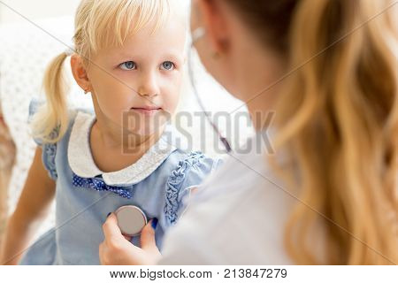 Serious face of little Caucasian girl visiting doctor. Female doctor examining girl with stethoscope. Pediatrics and health concept