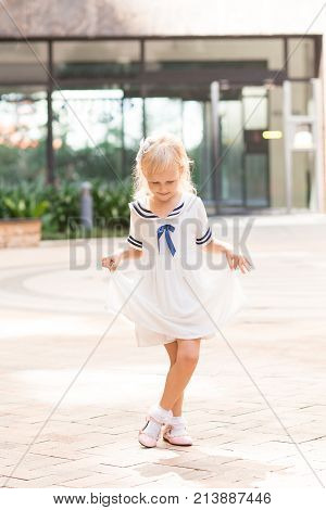 Portrait of pretty little fair-haired girl curtseying outdoors. Front view.