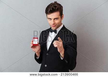 Portrait of a handsome charming man dressed in tuxedo holding open box with an engagement ring and pointing finger at camera isolated over gray background