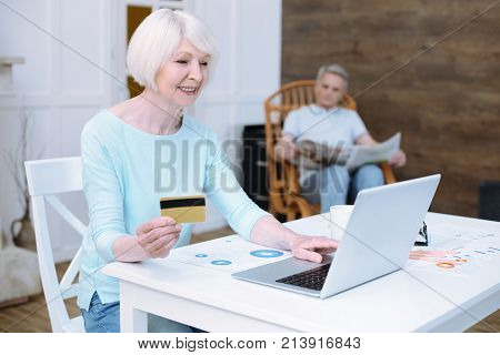 Financial moments. Smart progressive old woman using her credit card while paying the bills