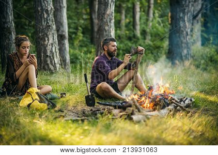 Couple of hikers relax at bonfire in forest. Man hipster sharpen axe. Girl or woman use mobile phone. New technology or archaism concept. Camping hiking summer vacation.