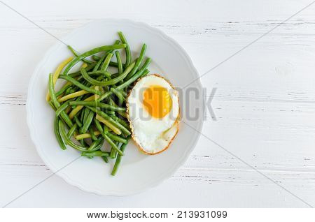 Cooked green beans with fried egg in white plate on wooden background with space for text. Healthy vegetarian food concept. Top view. Copy space.
