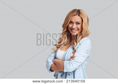 Beauty and style. Attractive young smiling woman keeping arms crossed and looking at camera while standing against grey background
