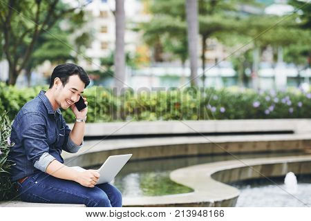 Cheerful young businessman calling on the phone and reading information on digital tablet