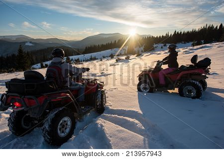 Young couple riders sitting on off-road quad bikes enjoying beautiful sunset in the the mountains in winter