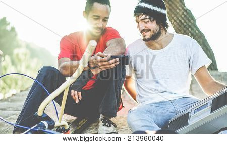 Bikers friends watching videos feed of their performances in city park outdoor - Young people having fun sharing media online of extreme sport - Focus on left man hand phone - Friendship concept