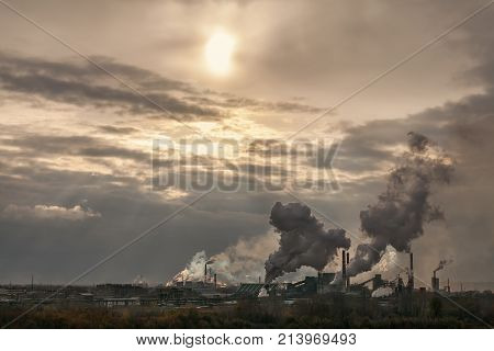 large plant of a lot of black smoke from the pipes against the background of a stormy sky the sun shines through the clouds with the rays of light