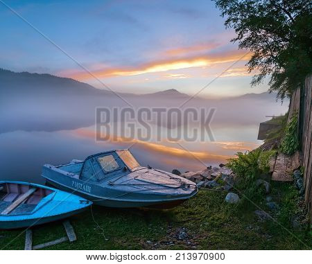 summer early morning on the lake in the fog. against the background of the mountains and the village on the shore. two blue fishing boats lie on the shore against a background of pink dawn