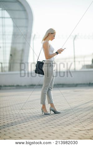 Attractive business woman using a phone and clock while standing in front of windows in an office building overlooking the city