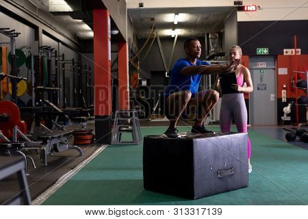 Side view of an African-American man doing plyometrics and a Caucasian woman assisting him inside a room at a sports centre