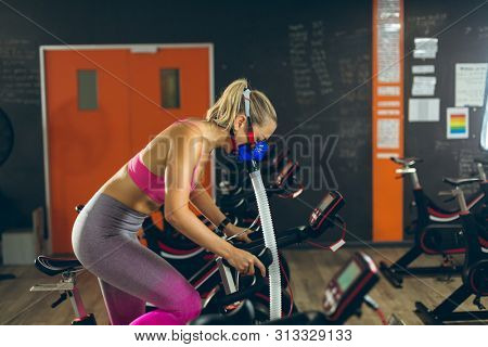 Side view of beautiful young Caucasian female athlete with oxygen mask exercising with exercise bike in fitness studio. Bright modern gym with fit healthy people working out and training