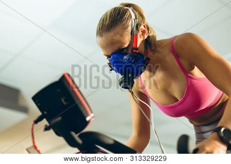 Low angle view of beautiful young Caucasian female athlete with oxygen mask exercising with exercise bike in fitness studio. Bright modern gym with fit healthy people working out and training