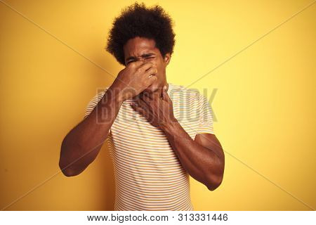 American man with afro hair wearing striped t-shirt standing over isolated yellow background smelling something stinky and disgusting, intolerable smell, holding breath with fingers on nose. Bad smel