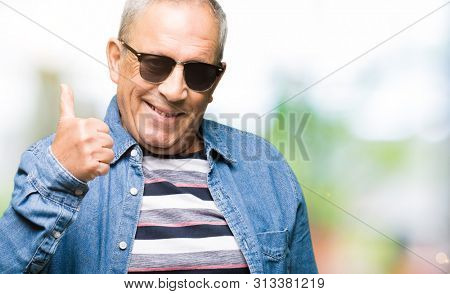 Handsome senior man wearing denim jacket and sunglasses doing happy thumbs up gesture with hand. Approving expression looking at the camera with showing success.