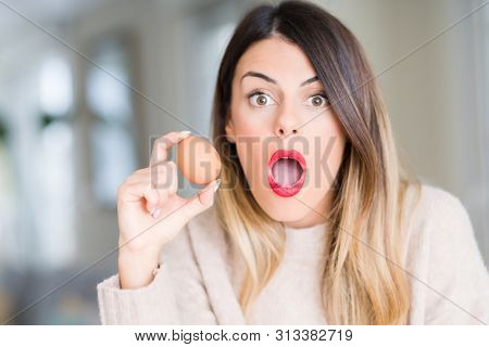 Young beautiful woman holding fresh egg at home scared in shock with a surprise face, afraid and excited with fear expression